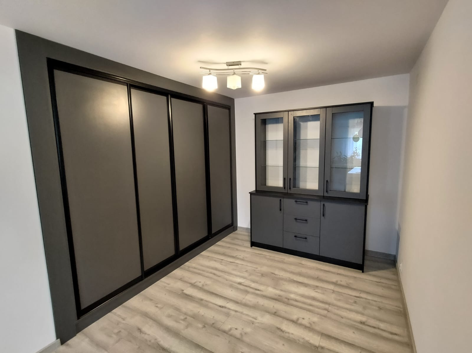 Szara szafa przesuwna z czterema drzwiami oraz szary kredens z przeszkloną nadstawką w minimalistycznym wnętrzu z podłogą imitującą drewno.