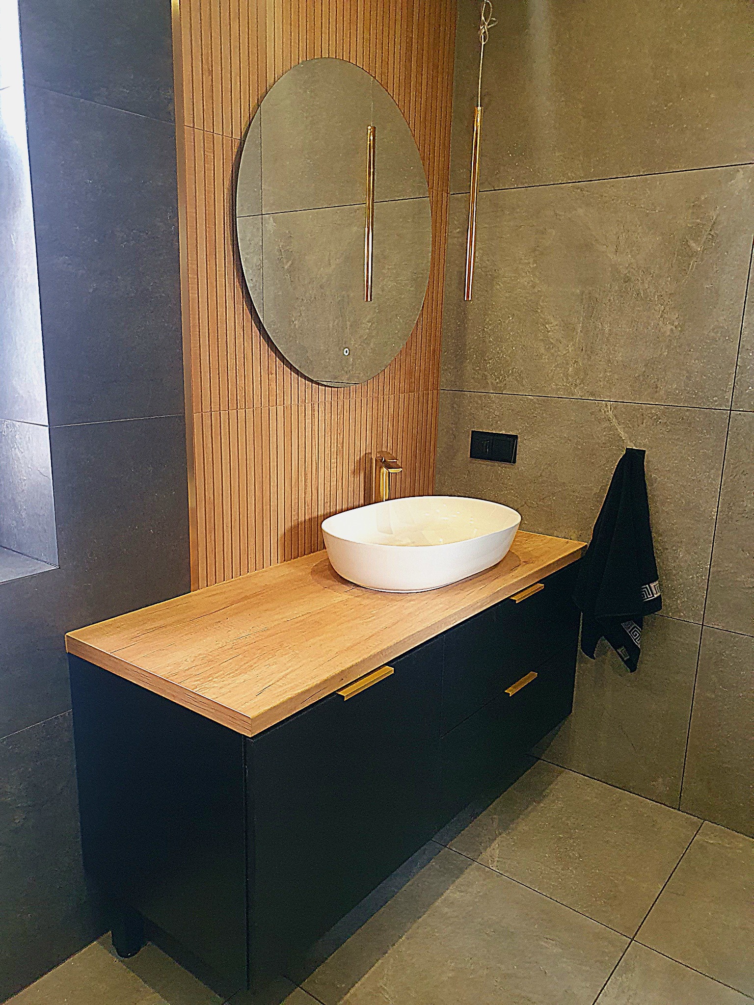 Nowoczesna łazienka z czarną szafką pod umywalkę z drewnianym blatem, owalną białą umywalką nablatową, złotą armaturą i okrągłym lustrem z podświetleniem na tle drewnianej ściany lamelowej...