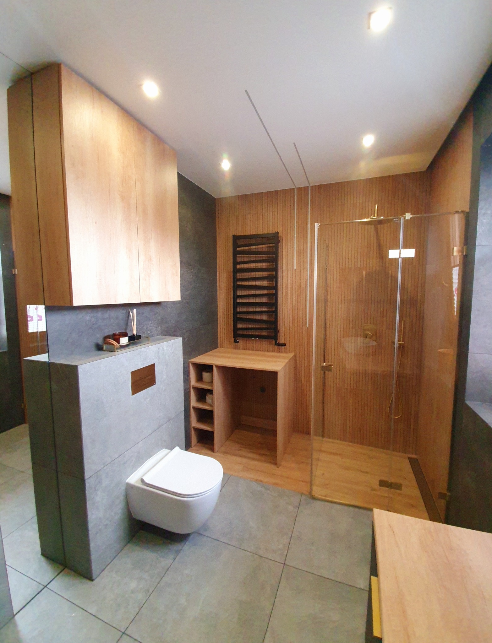 Nowoczesna łazienka z drewnianymi akcentami: wisząca toaleta, szklana kabina prysznicowa, drewniana szafka z półkami i grzejnik drabinkowy na tle drewnianej ściany.