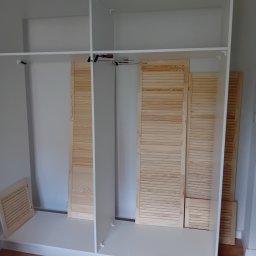 Łukasz Kalinowski - Wnętrze szafy wnękowej w trakcie montażu, widoczne białe półki i drewniane żaluzjowe drzwi, na podłodze leży wkrętarka.