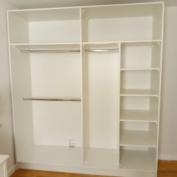 Łukasz Kalinowski - Wnętrze białej, modułowej garderoby z drążkami i półkami, widoczny fragment drewnianej podłogi i dywanu.