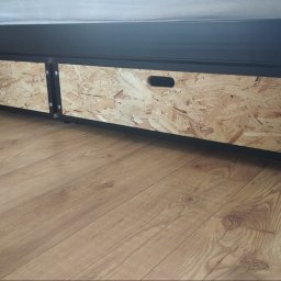 Łukasz Kalinowski - Dwie szuflady na kółkach z płyty OSB, umieszczone pod czarnym łóżkiem, widoczne na tle podłogi z paneli imitujących drewno.