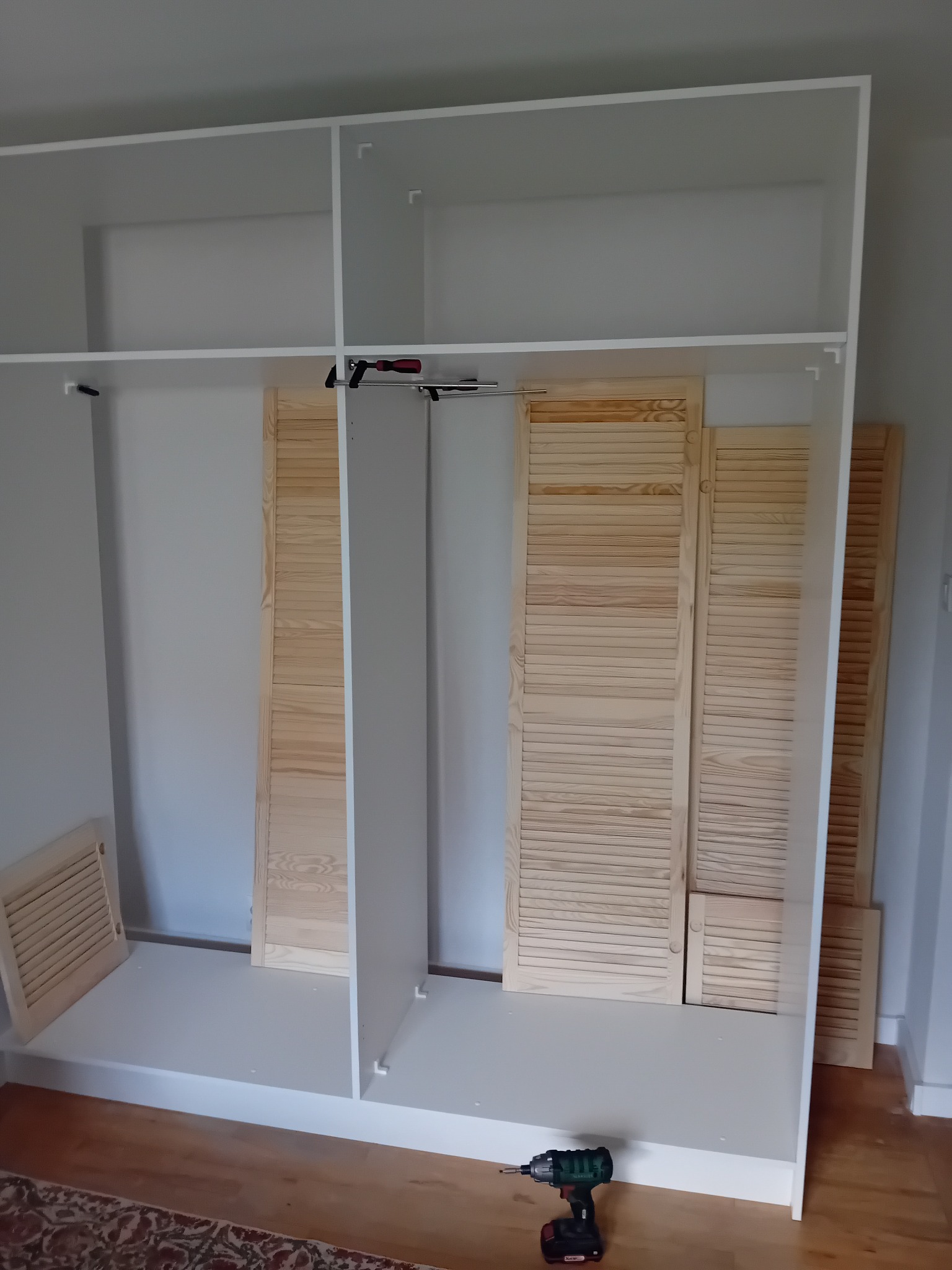 Wnętrze szafy wnękowej w trakcie montażu, widoczne białe półki i drewniane żaluzjowe drzwi, na podłodze leży wkrętarka.