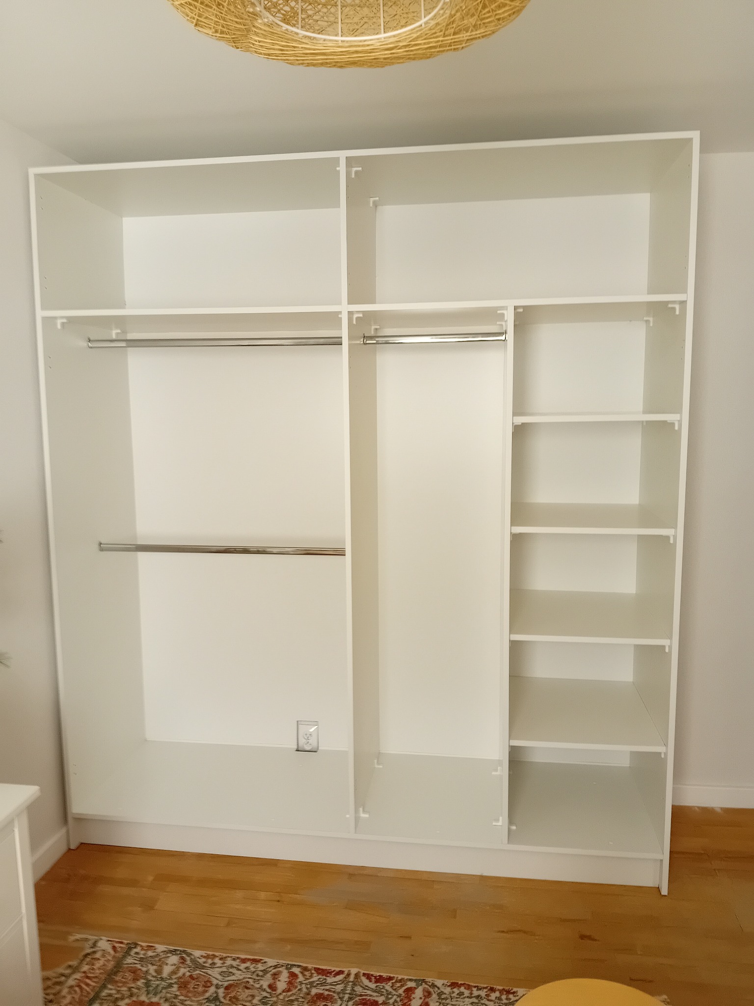 Wnętrze białej, modułowej garderoby z drążkami i półkami, widoczny fragment drewnianej podłogi i dywanu.
