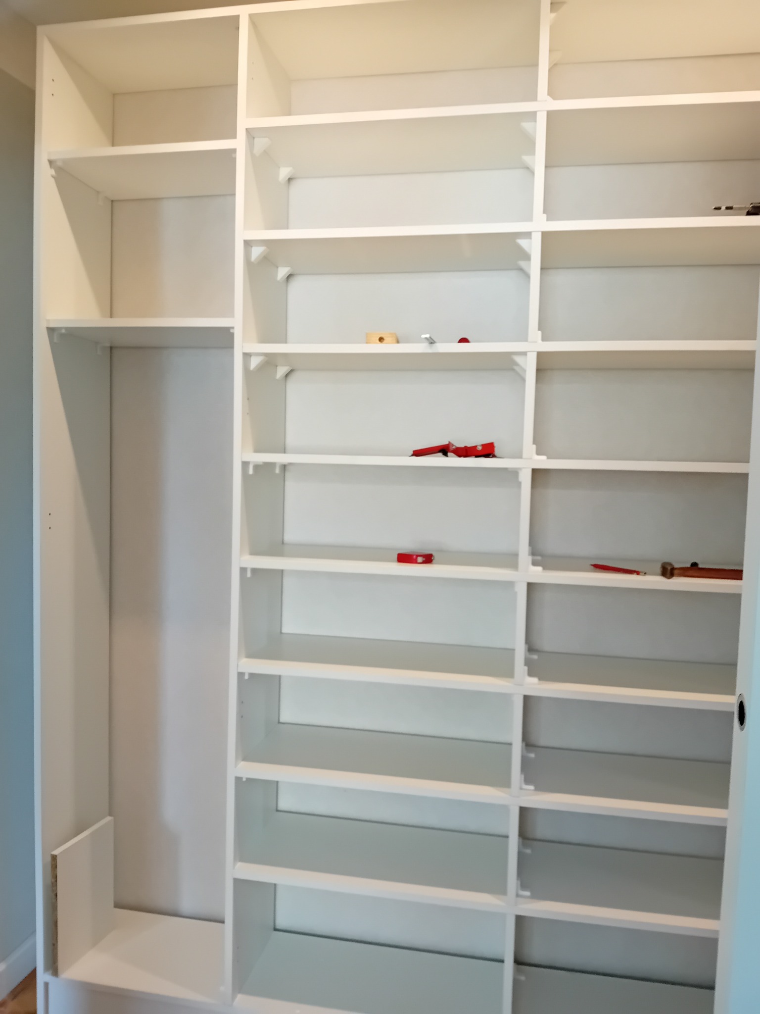 Wnętrze białej, modułowej szafy z otwartymi półkami i miejscem na wieszaki, widoczne narzędzia na półkach, sugerujące montaż lub regulację.