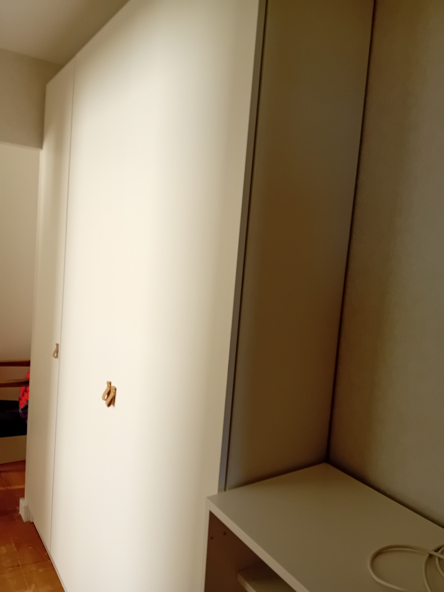 Wysoka, biała szafa z trzema drzwiami i skórzanymi uchwytami, ustawiona w rogu pokoju, obok jasnej półki z widocznym kablem.