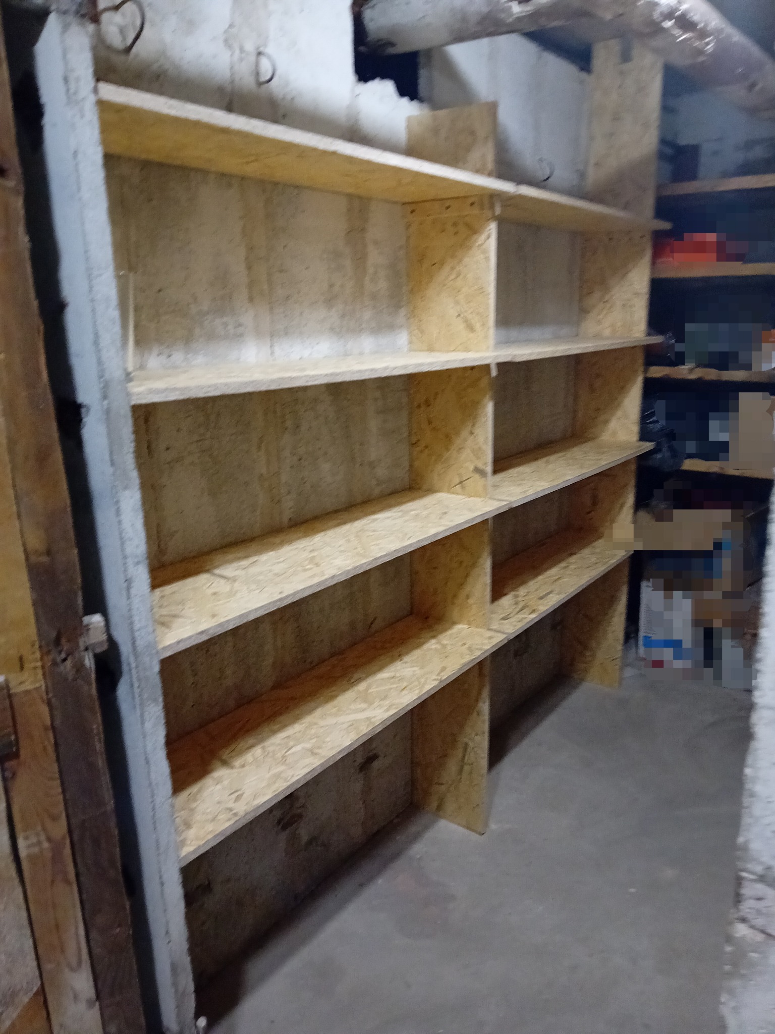 Surowe, drewniane regały z płyty OSB w piwnicy, z widocznymi elementami starej konstrukcji budynku i rurą pod sufitem.