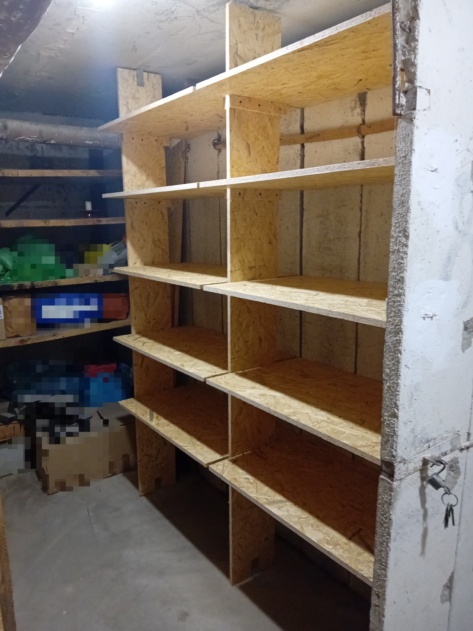 Surowe, drewniane półki z płyty OSB w piwnicy, z widocznymi elementami mocującymi i betonową ścianą obok, dodatkowe półki w tle.
