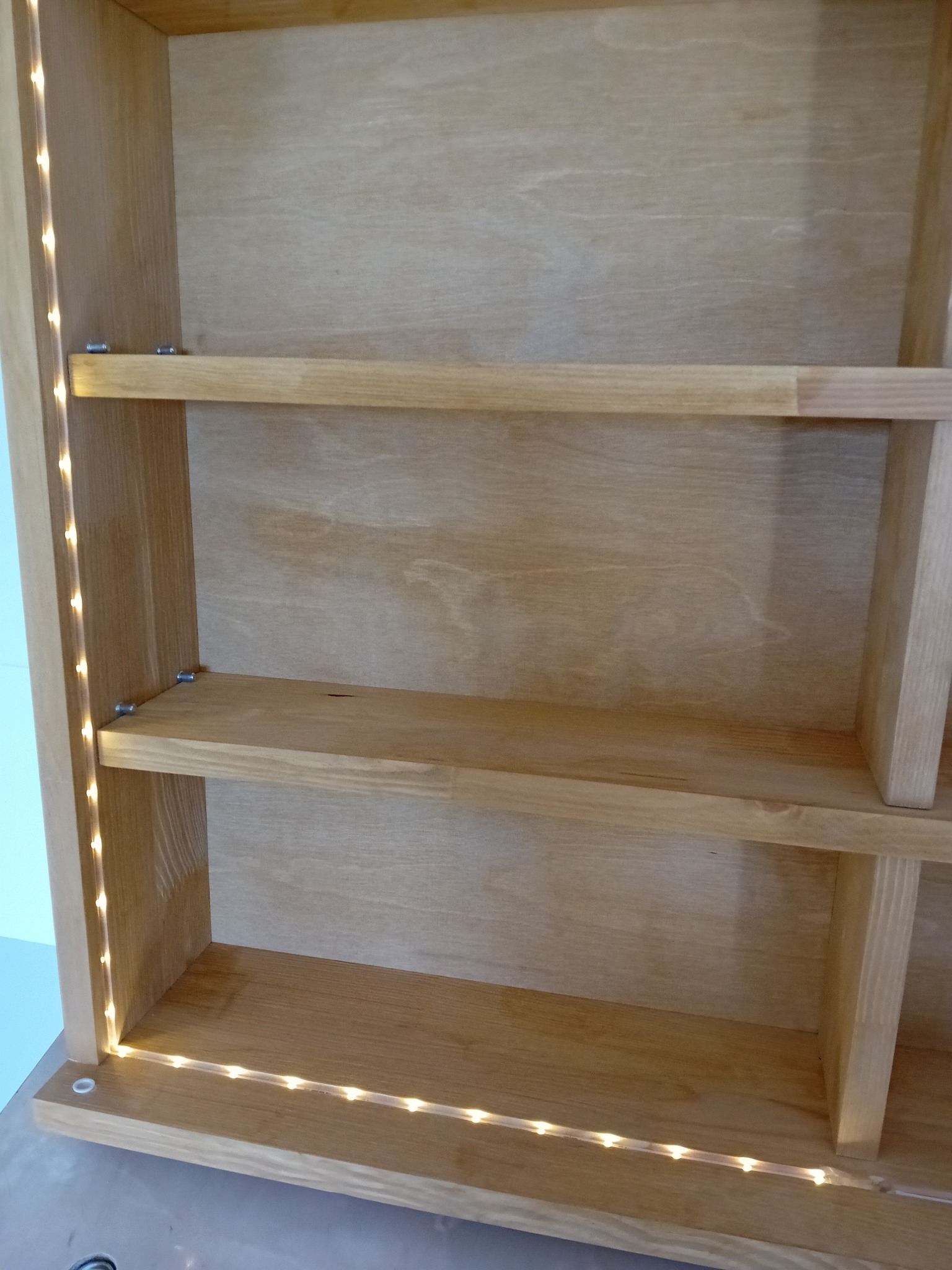 Wnętrze drewnianej szafki z półkami, oświetlone listwą LED wzdłuż krawędzi bocznej ściany, z widocznymi mocowaniami półek.
