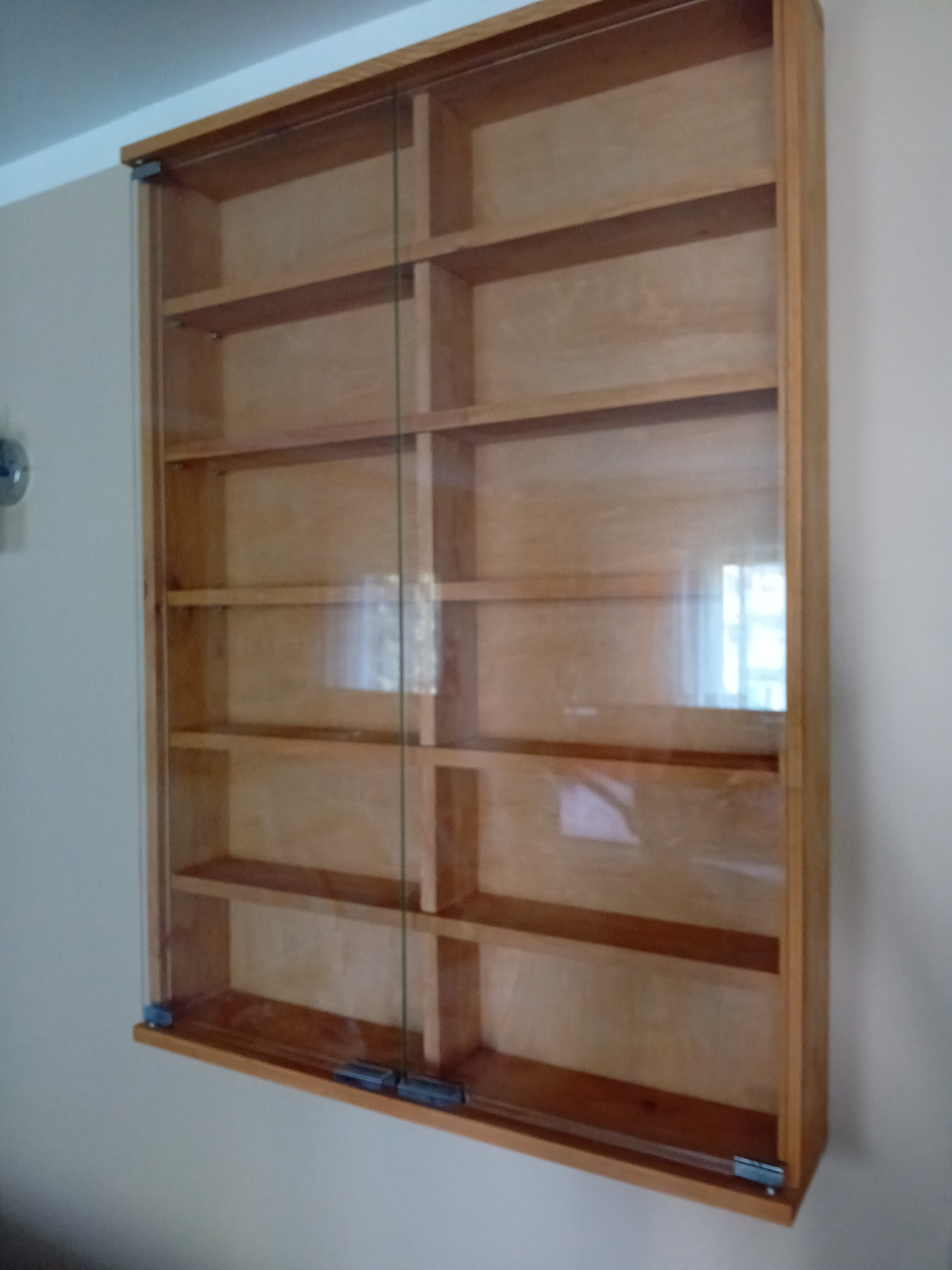 Drewniana szafka wisząca z przesuwnymi szklanymi drzwiami, puste półki, widoczne mocowania do ściany.