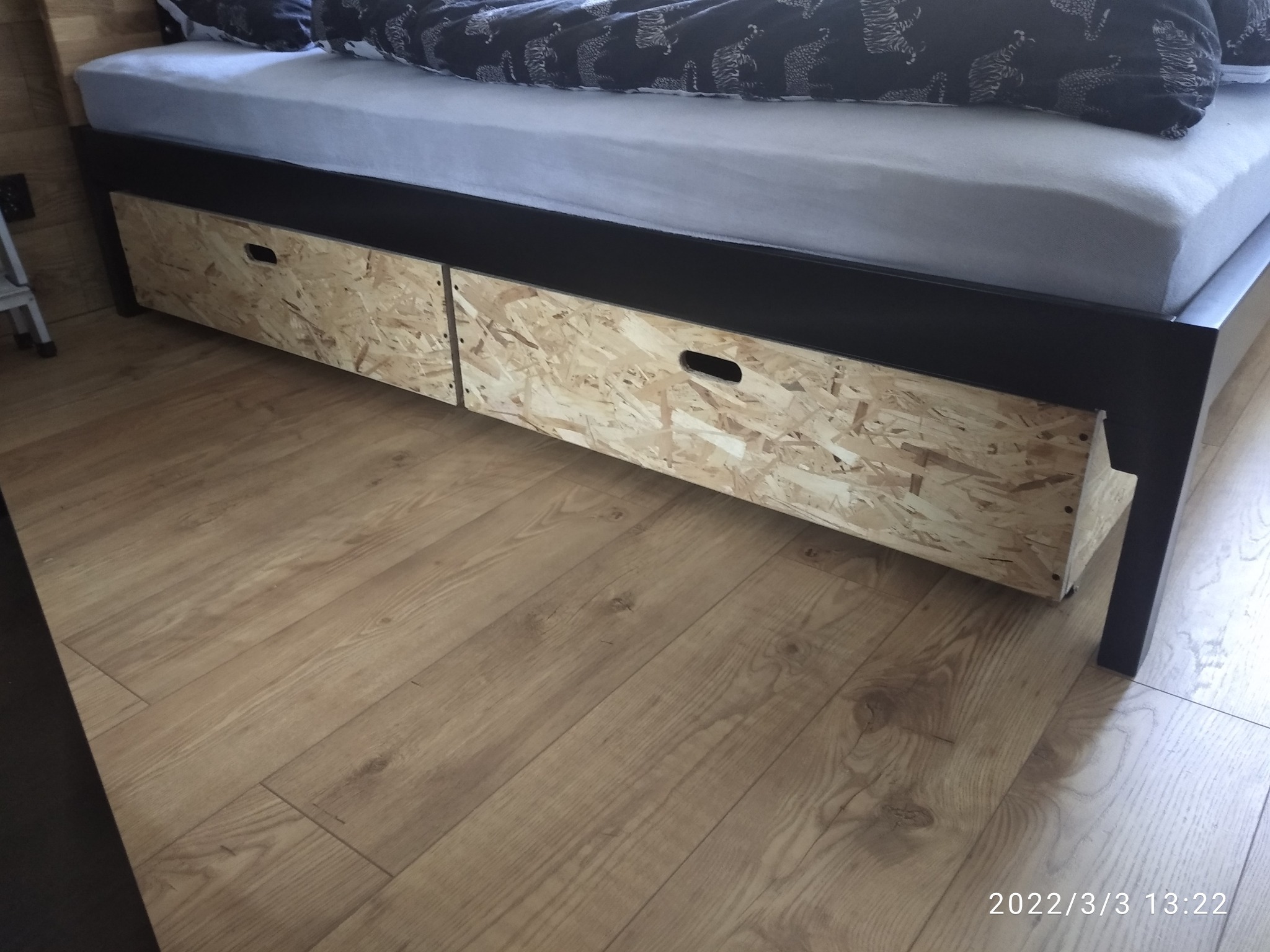Łóżko z czarną ramą i dwiema szufladami z płyty OSB na kółkach, umieszczonymi pod spodem, na podłodze z paneli.