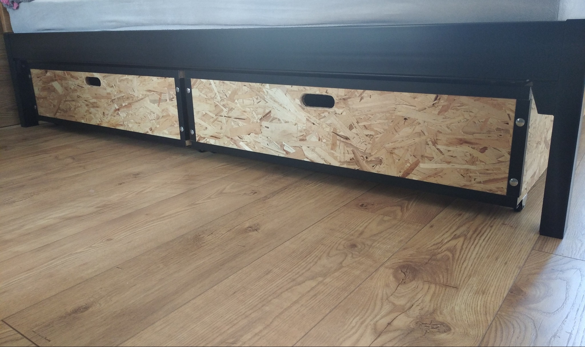 Dwie szuflady na kółkach z płyty OSB, umieszczone pod czarnym łóżkiem, widoczne na tle podłogi z paneli imitujących drewno.