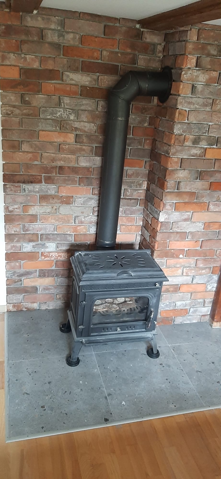 Żeliwny piec wolnostojący z czarną rurą odprowadzającą spaliny, ustawiony na szarych płytkach, z tłem ściany wyłożonej cegłą w odcieniach czerwieni i brązu.