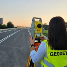 Kobieta w kamizelce odblaskowej z napisem, obsługuje tachimetr na tle nowej drogi, widok z tyłu, w oddali słup wysokiego napięcia i góry.