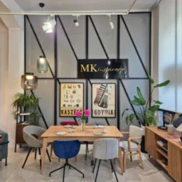 Jadalnia z drewnianym stołem, r&oacute;żnymi krzesłami i dywanem w jodełkę. W tle ściana z metalową kratą i plakatami. Lampy wiszące i szafka z rośliną.