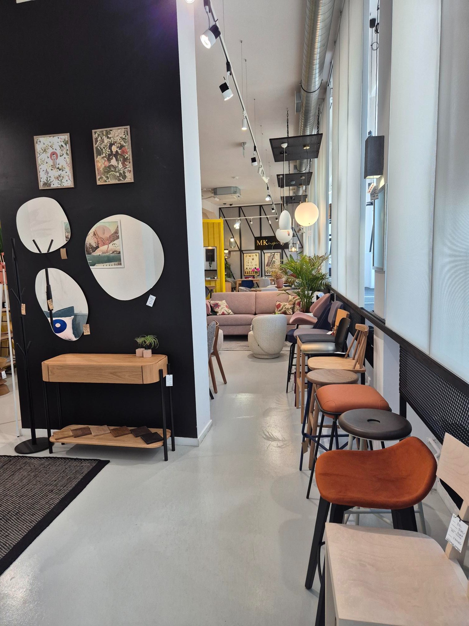 Wnętrze salonu meblowego z ekspozycją krzeseł barowych, stolika z lustrami i dekoracjami na czarnej ścianie oraz jasnej sofy w głębi. Oświetlenie punktowe.