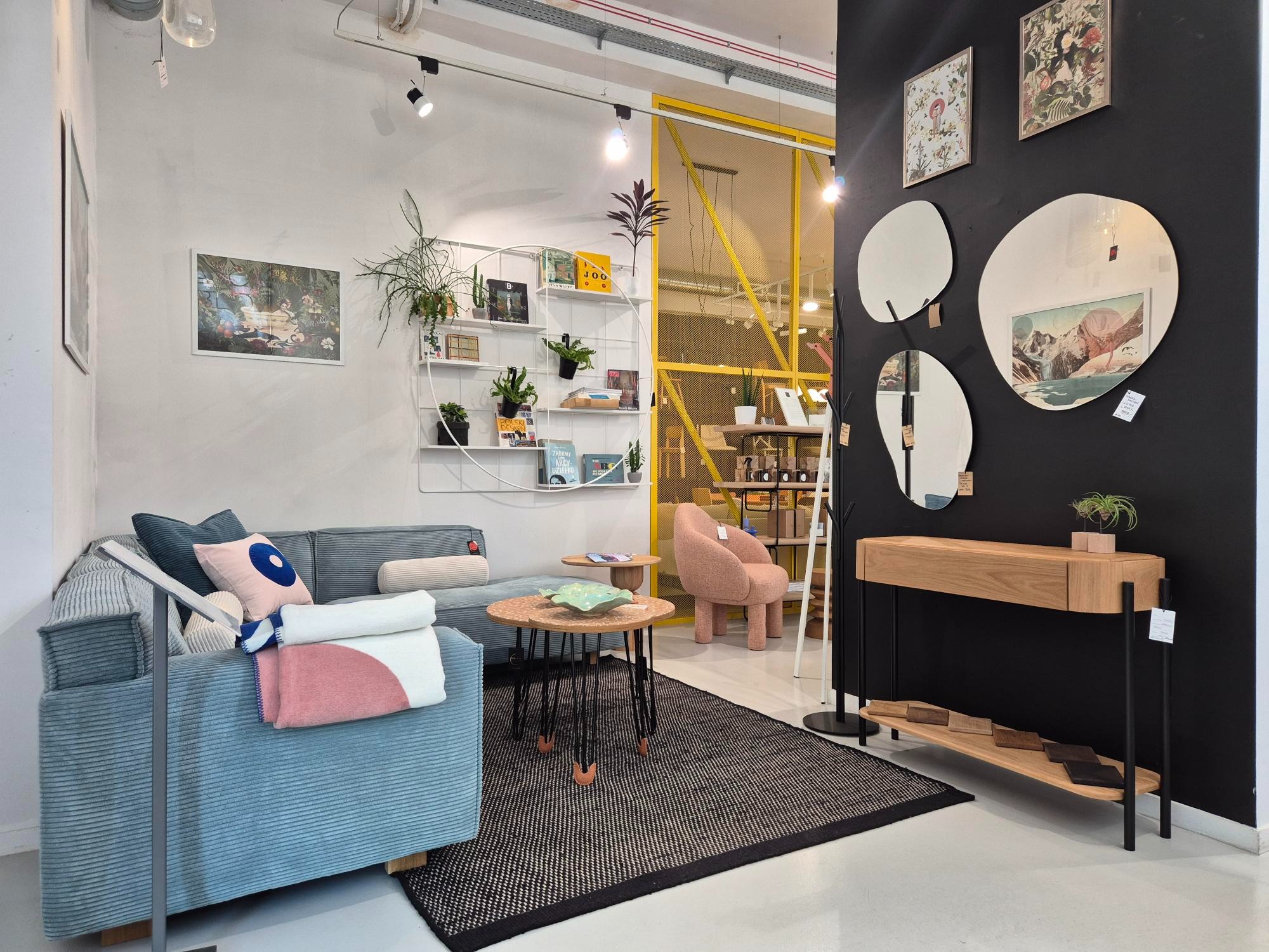 Stylowe wnętrze: błękitna sofa z poduszkami, okrągłe stoliki kawowe, czarny dywan, konsola z drewna i lustra na czarnej ścianie. Elementy dekoracyjne i rośliny.