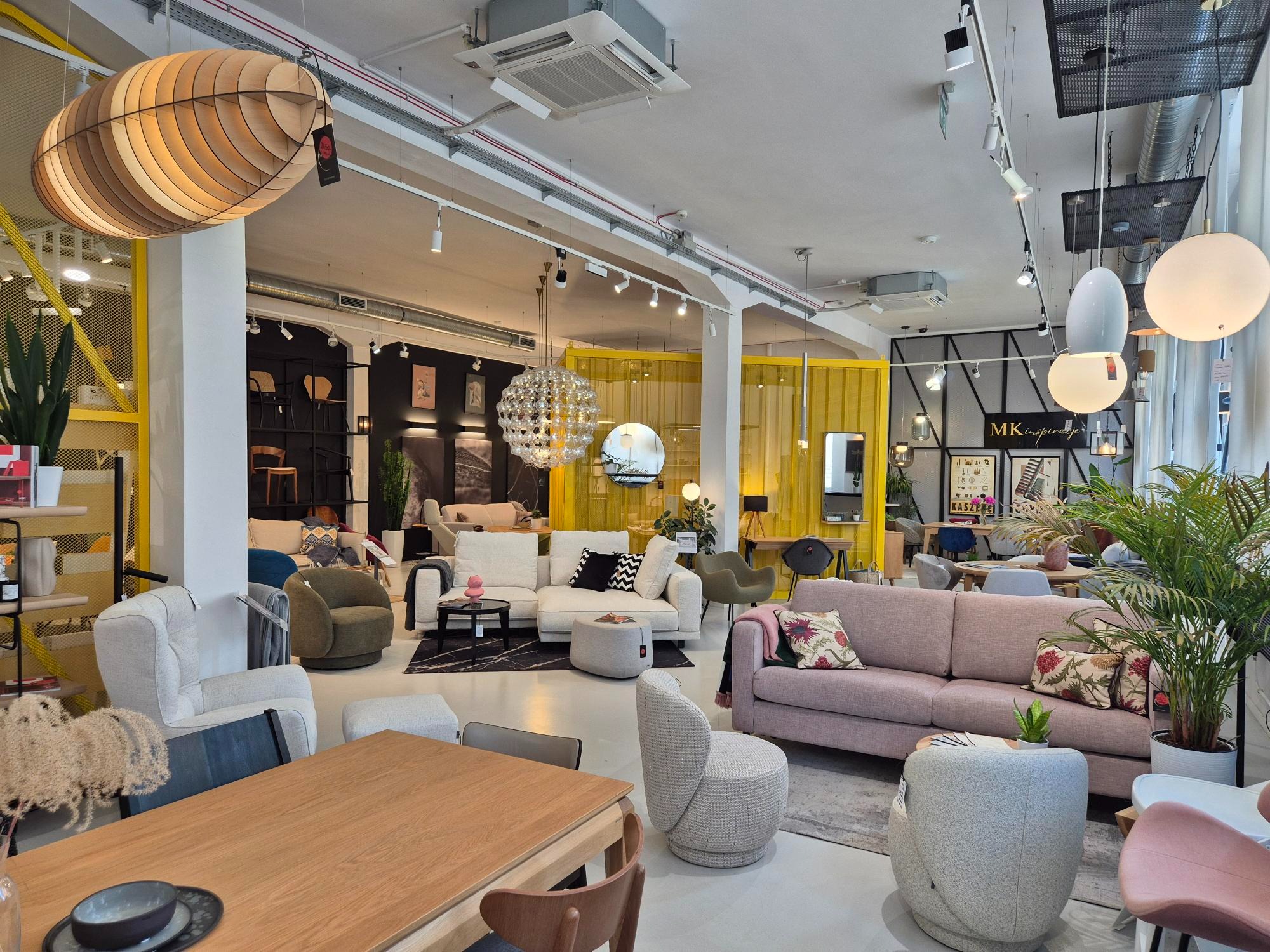 Salon meblowy z ekspozycją sof, foteli i stołów w różnych stylach, oświetlony designerskimi lampami, z roślinami doniczkowymi, tworzący przytulną atmosferę.