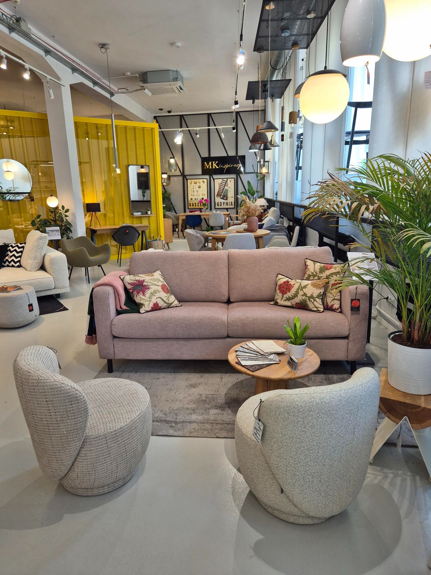 Salon meblowy: różowa sofa z poduszkami w kwiaty, dwa szare fotele, stolik kawowy, roślina doniczkowa. W tle ekspozycja z żółtymi elementami i inne meble.