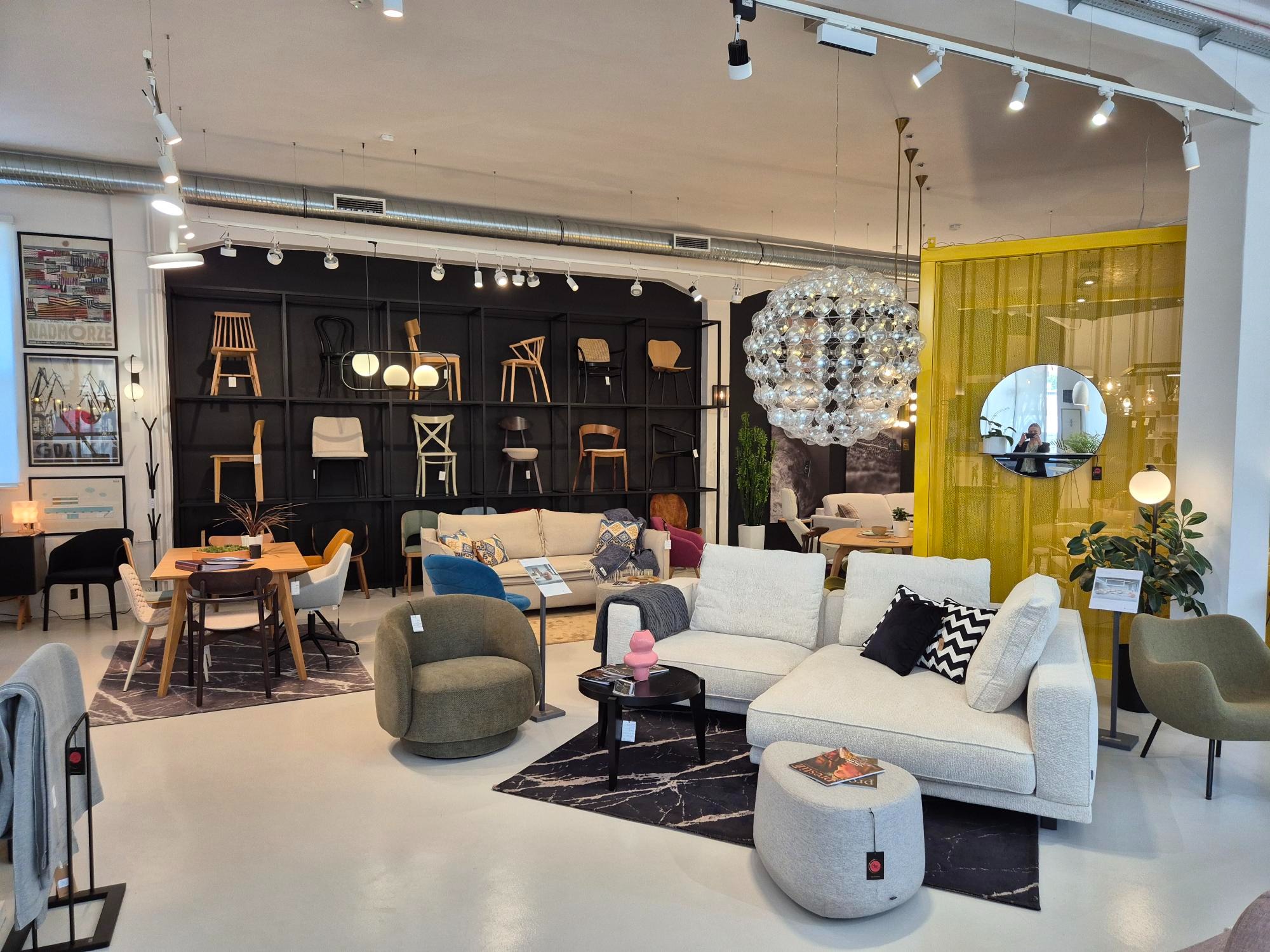 Salon meblowy z ekspozycją różnorodnych mebli: sofy modułowe, krzesła, stoły i lampy. W tle ściana z krzesłami. Nowoczesny design, jasne wnętrze.