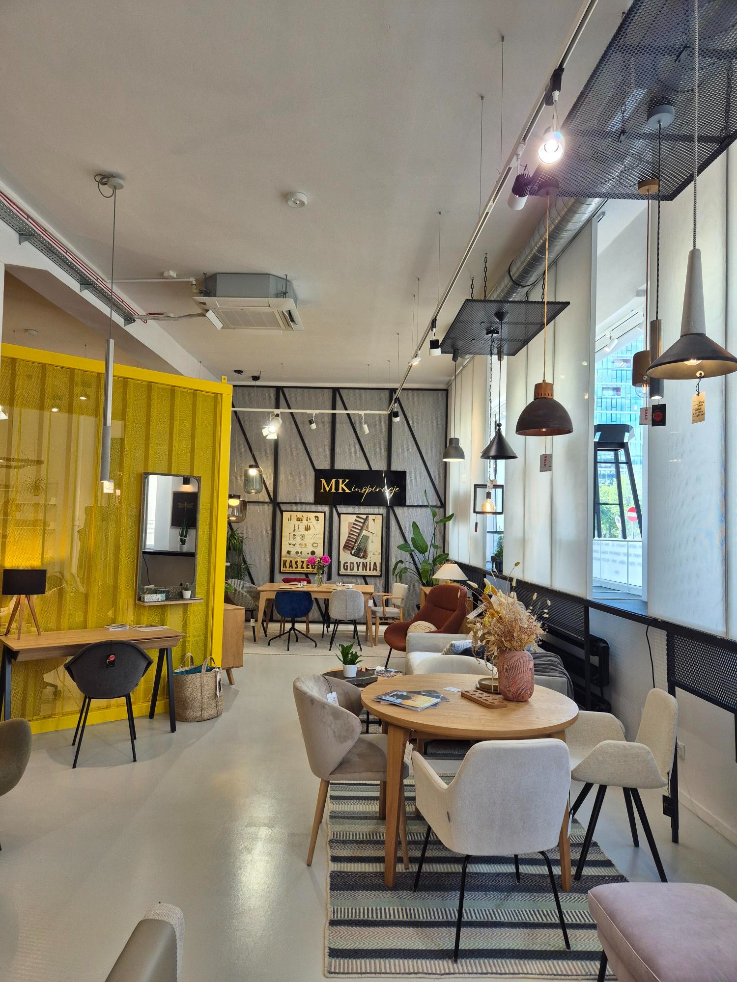 Jasne wnętrze salonu meblowego z ekspozycją krzeseł tapicerowanych, stołów i dekoracji. Żółty parawan oddziela przestrzeń biurową. Lampy wiszące nadają nowoczesny charakter.