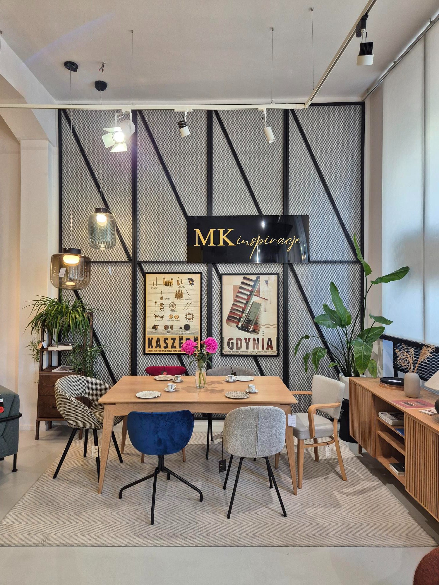 Jadalnia z drewnianym stołem, różnymi krzesłami i dywanem w jodełkę. W tle ściana z metalową kratą i plakatami. Lampy wiszące i szafka z rośliną.