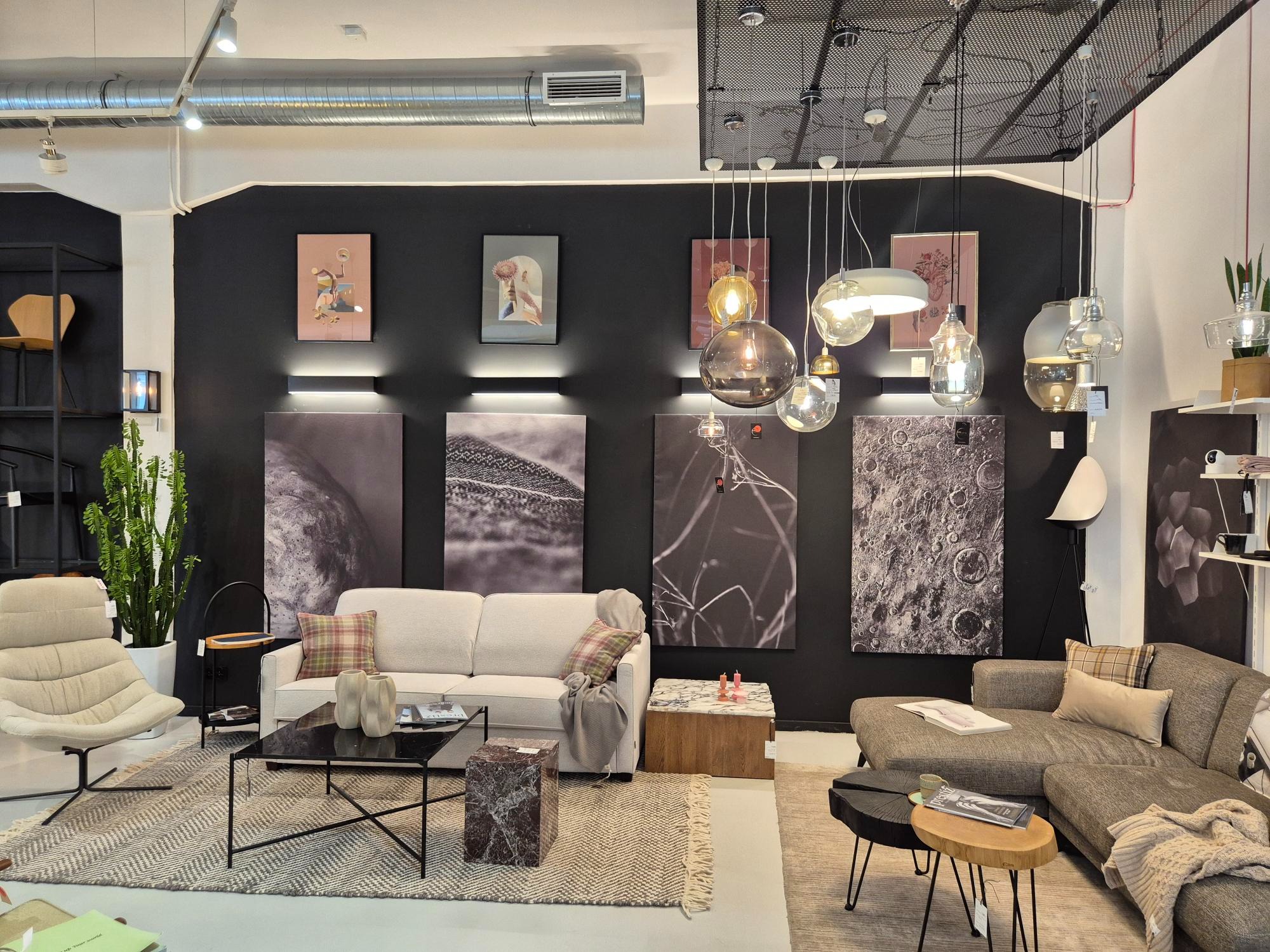 Salon z sofą w neutralnych barwach, czarnym tłem z grafikami i lampami. Widoczne stoliki kawowe i dywany w stylu skandynawskim, tworzące przytulną przestrzeń.