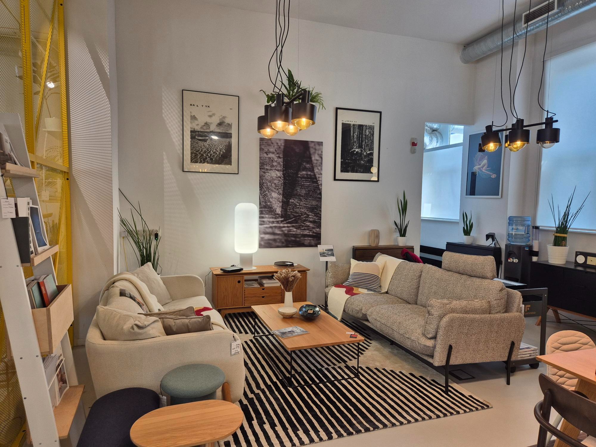 Jasne wnętrze salonu z dwoma sofami, stolikiem kawowym na czarno-białym dywanie w paski. Nad nimi wiszą lampy z roślinami. Na ścianach grafiki w czarnych ramkach.