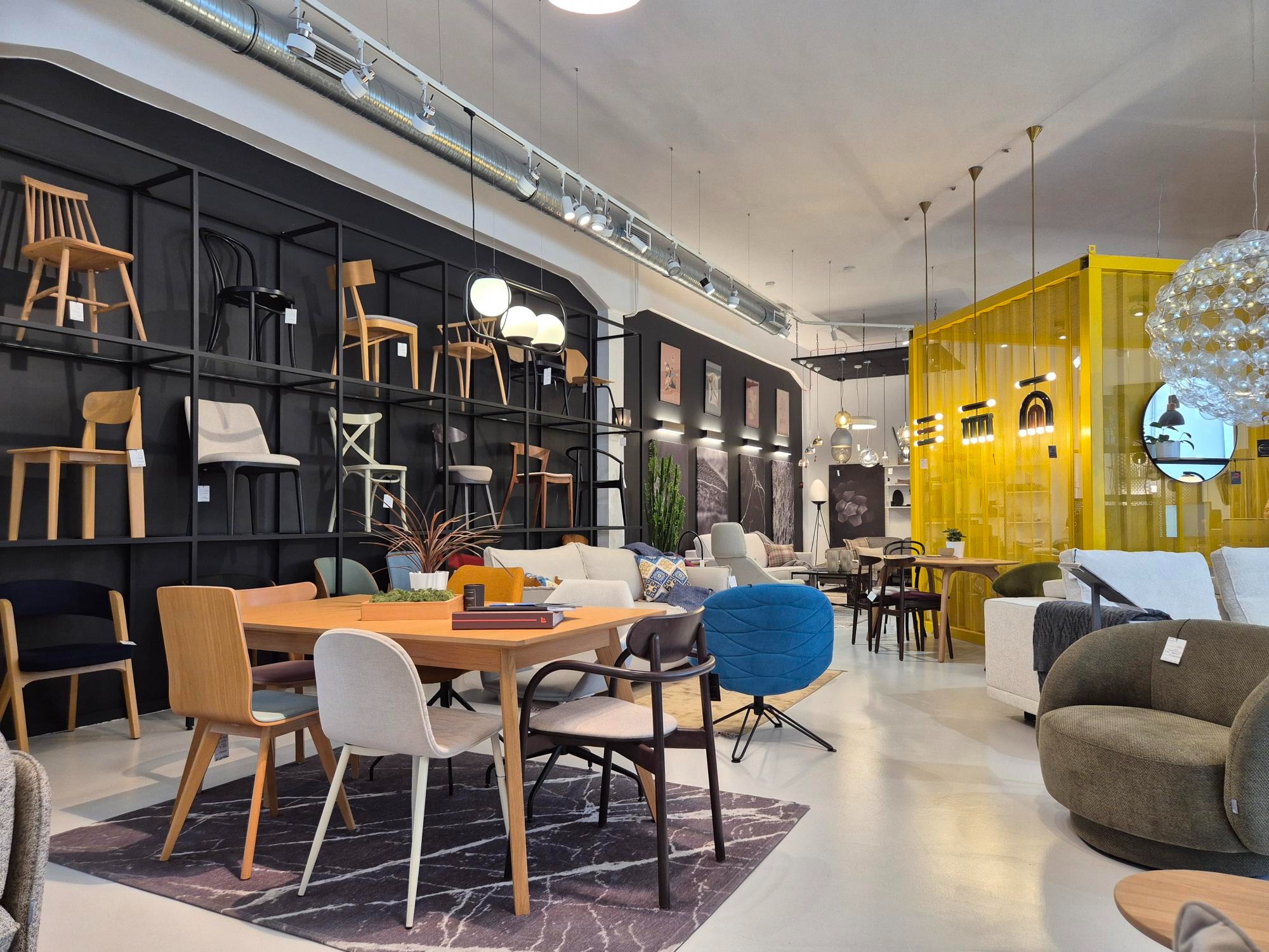 Salon meblowy z ekspozycją krzeseł na ścianie, stołu jadalnego i tapicerowanych foteli w różnych stylach, oświetlony lampami wiszącymi.