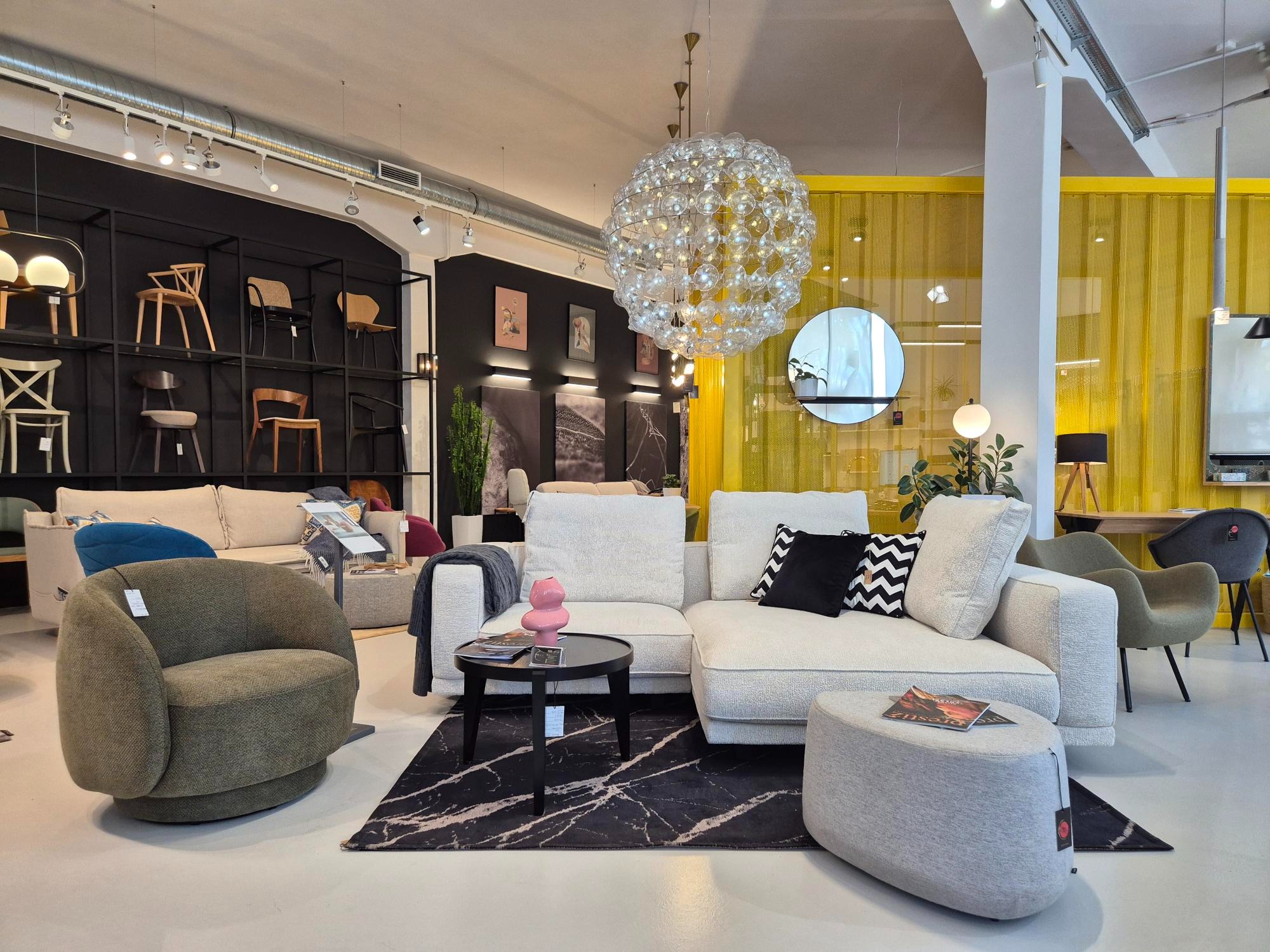 Jasny narożnik tapicerowany w salonie meblowym, z okrągłym fotelem i czarnym stolikiem na tle czarnej ściany z ekspozycją krzeseł i żółtej dekoracji okiennej.