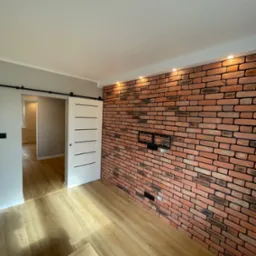 Pok&oacute;j po remoncie z ceglaną ścianą, przesuwnymi drzwiami i panelami na podłodze, widoczne pakiety paneli w rogu.