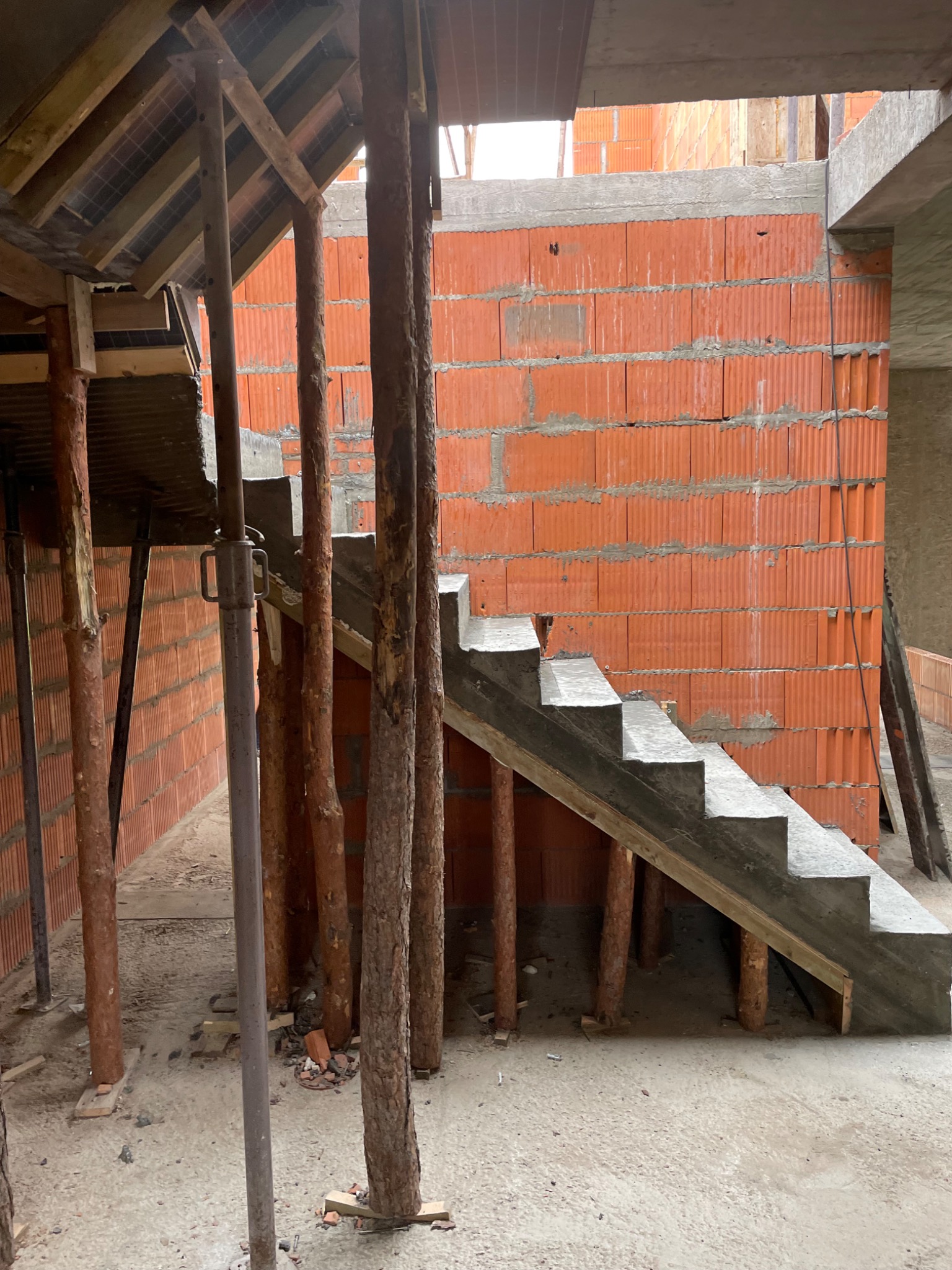 Wnętrze budynku w trakcie budowy z widocznymi betonowymi schodami wspartymi na drewnianych stemplach, ściany z czerwonej cegły dziurawki, surowy betonowy strop.