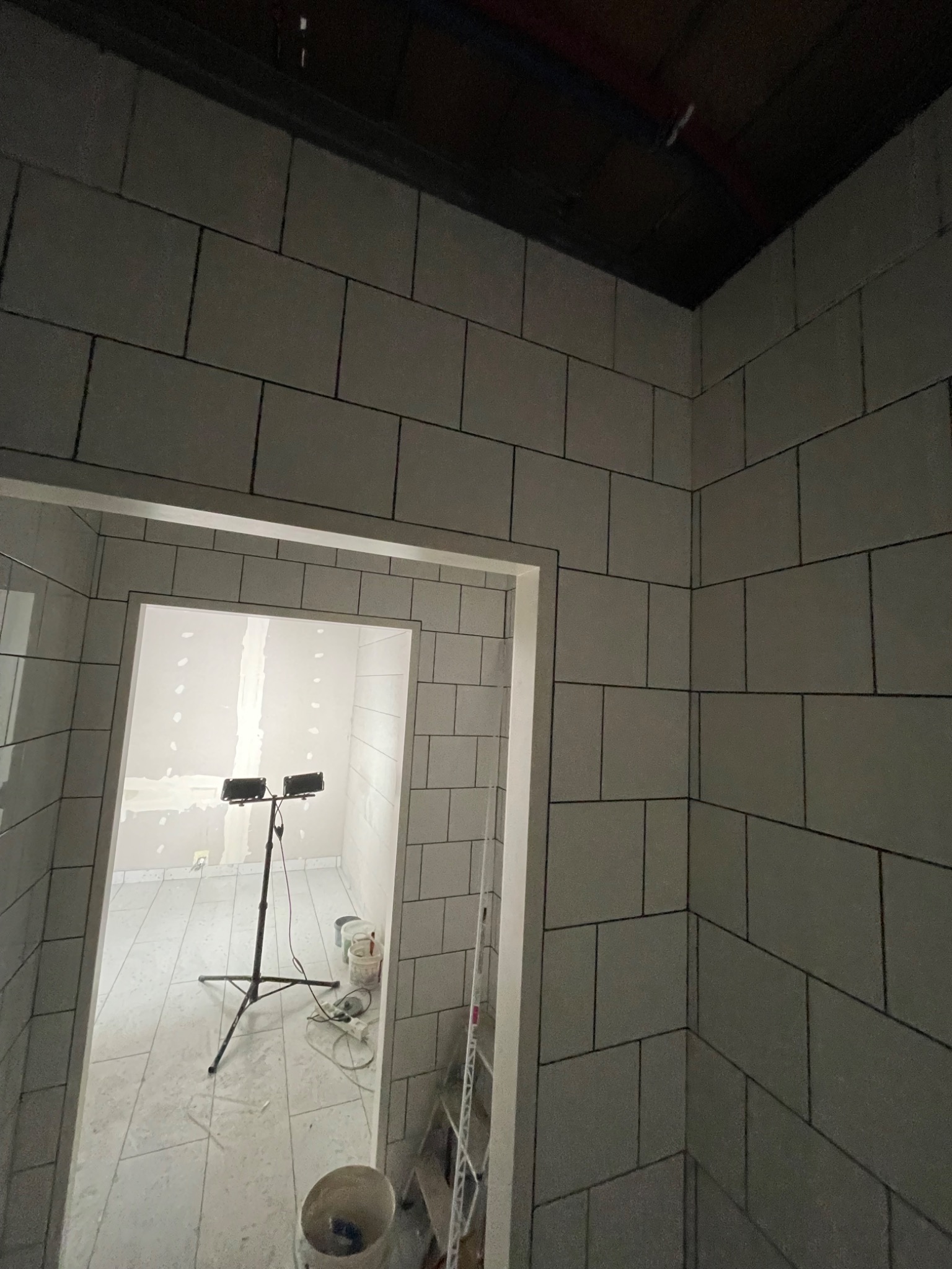 Świeżo wykafelkowana łazienka z szarymi płytkami i ciemną fugą, widoczne oświetlenie budowlane na statywie, wiadro i drabina w tle, sufit jeszcze nie wykończony.