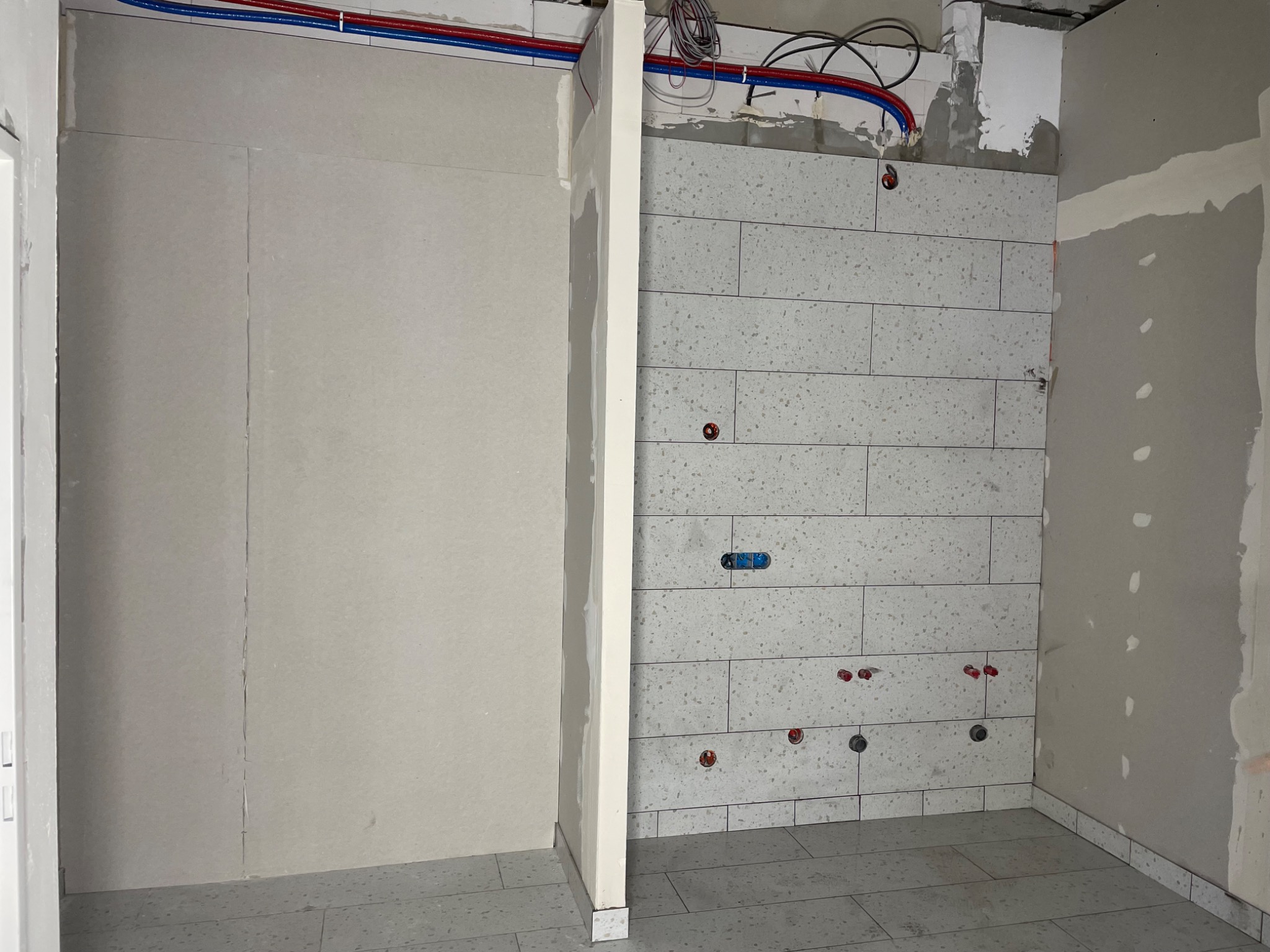 Łazienka w trakcie remontu: ułożone jasne, nakrapiane płytki na ścianie i podłodze, widoczne wyprowadzenia instalacji wodnej i elektrycznej, ściana przygotowana do malowania, surowe wykończenie.