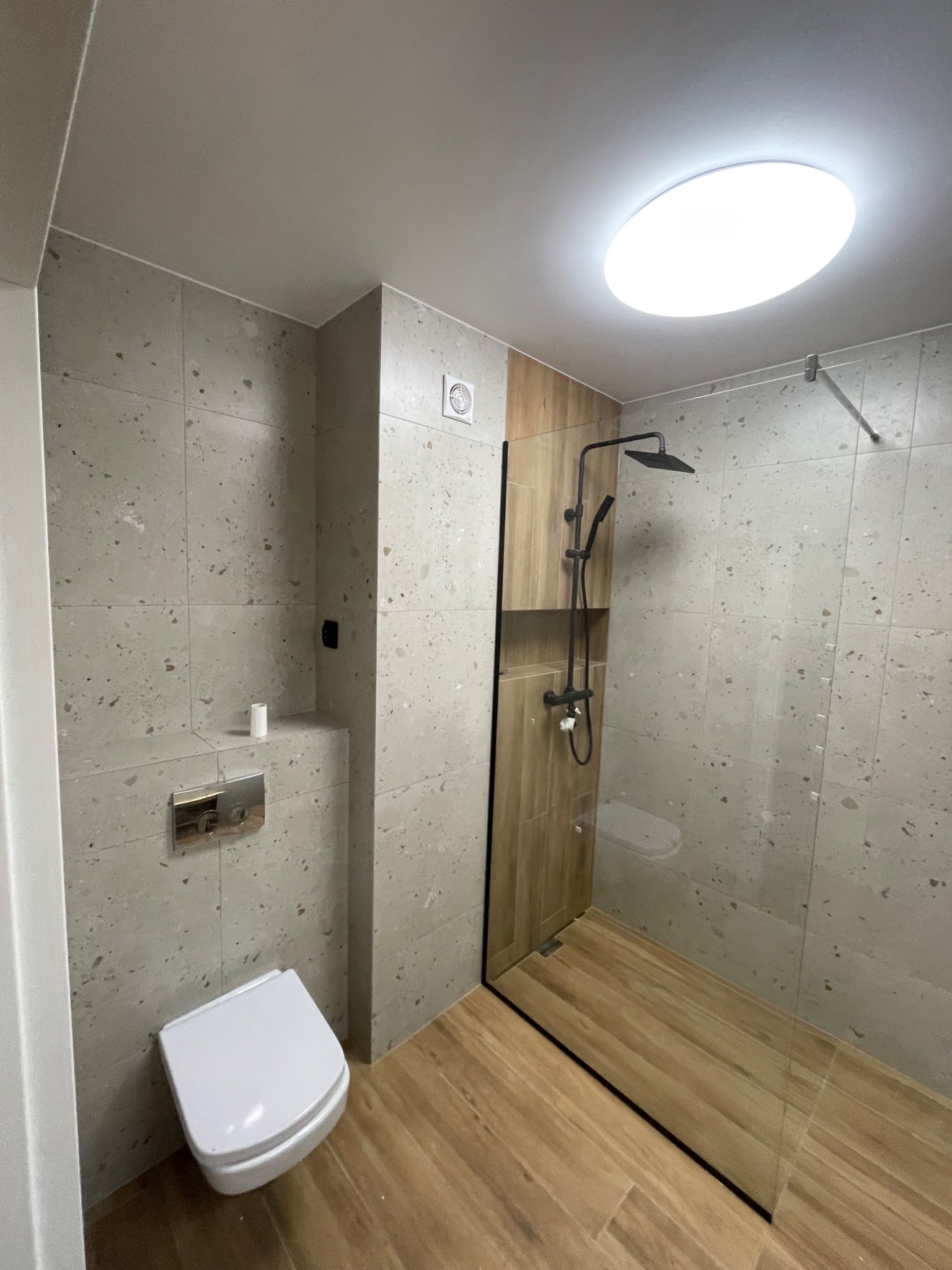 Nowoczesna łazienka z beżowymi płytkami imitującymi kamień, drewnopodobną podłogą w strefie prysznica walk-in z czarną deszczownicą i białą wiszącą toaletą.