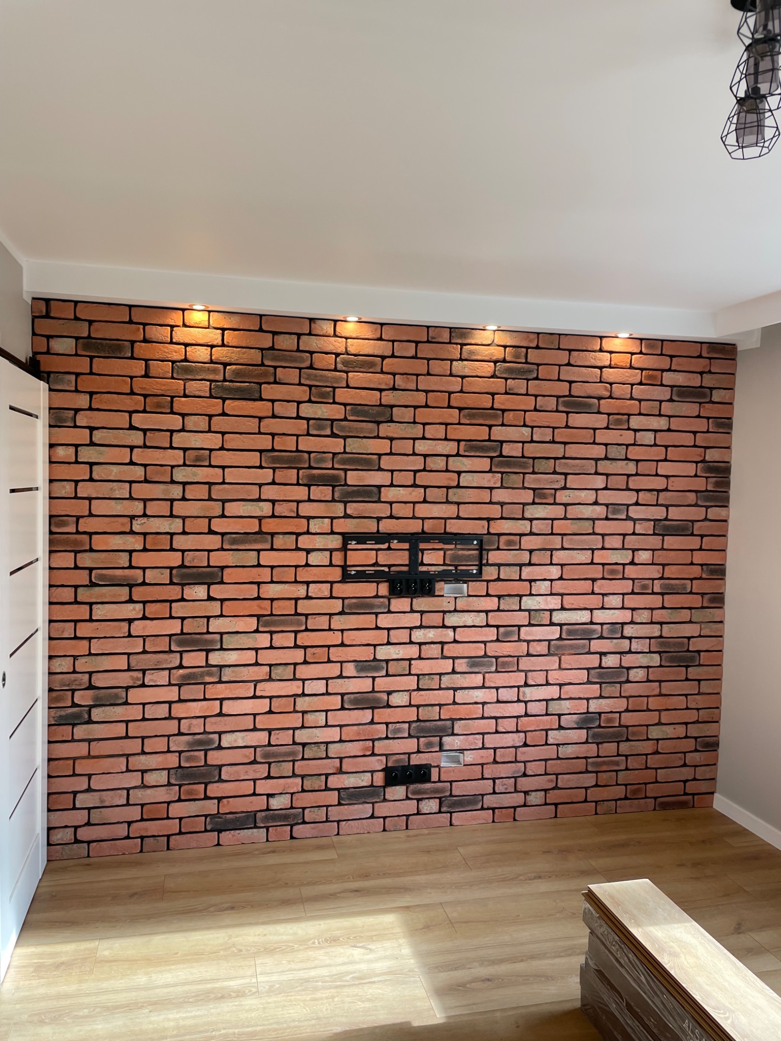 Ściana z cegły klinkierowej z zamocowanym uchwytem na telewizor i gniazdkami elektrycznymi, oświetlona punktowymi lampami sufitowymi, z ułożonymi panelami podłogowymi w pomieszczeniu.