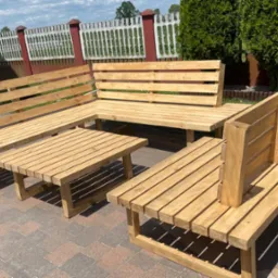 Zestaw mebli ogrodowych z jasnego drewna: narożna ławka, stół i pojedyncza ławka z wysokim oparciem, ustawione na kostce brukowej, w tle zielony trawnik i ogrodzenie.