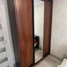 Brązowa szafa przesuwna z lustrem, czarne obramowanie, widoczna część pokoju w odbiciu lustra i fragment wzorzystej tapety na ścianie.