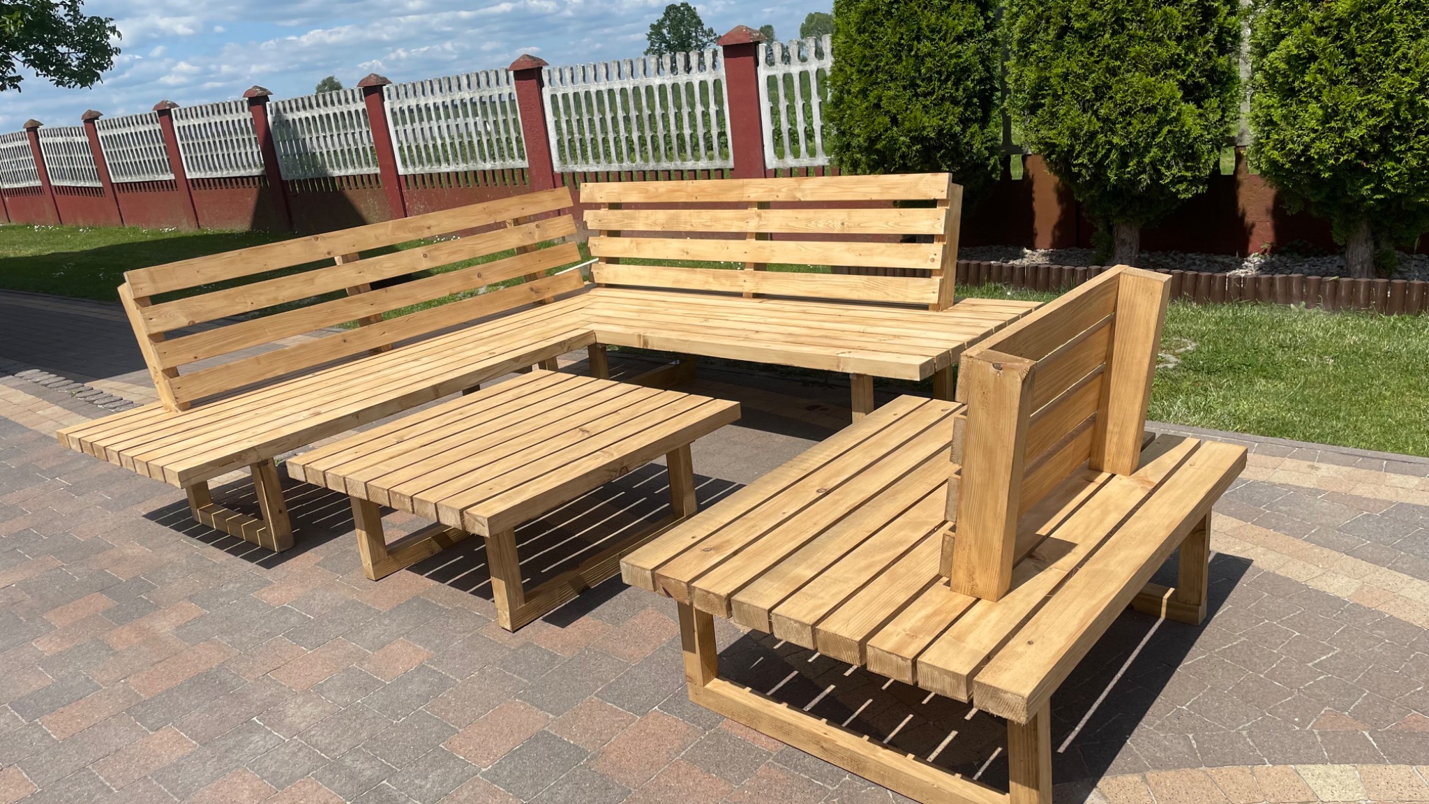 Zestaw mebli ogrodowych z jasnego drewna: narożna ławka, stół i pojedyncza ławka z wysokim oparciem, ustawione na kostce brukowej, w tle zielony trawnik i ogrodzenie.