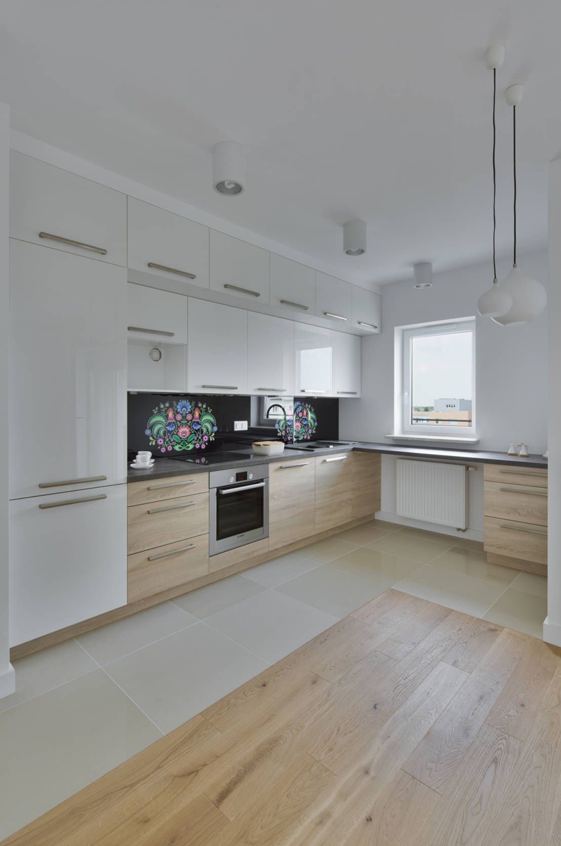 Jasna kuchnia z białymi szafkami wiszącymi i drewnopodobnymi szafkami dolnymi, czarnym blatem i folkowym motywem na ścianie nad blatem, oświetlona trzema lampami sufitowymi i dwoma wiszącymi lampami.