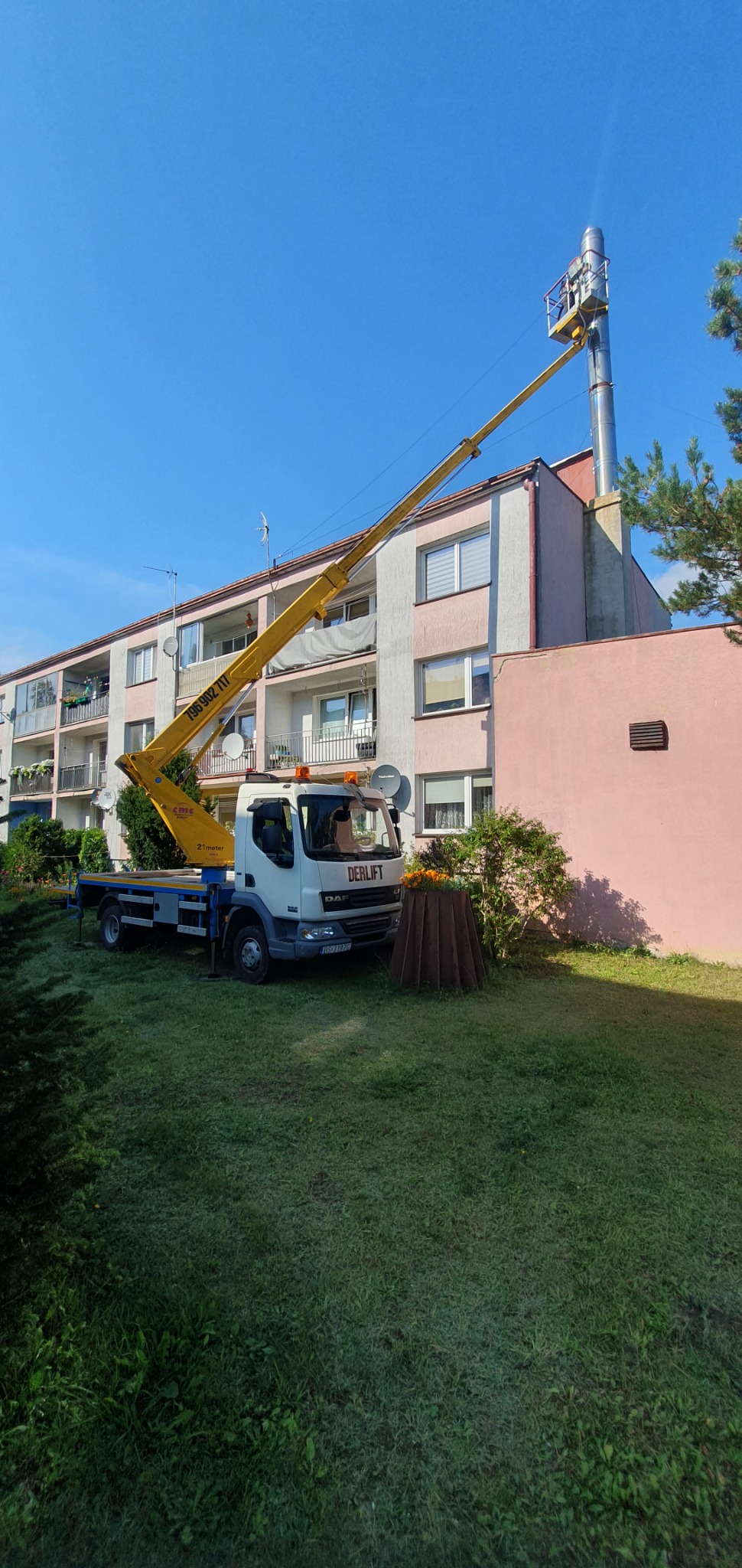Żółty pojazd z wysięgnikiem marki Derlift na trawniku, pracownik w koszu na szczycie komina na tle bloku mieszkalnego w słoneczny dzień.