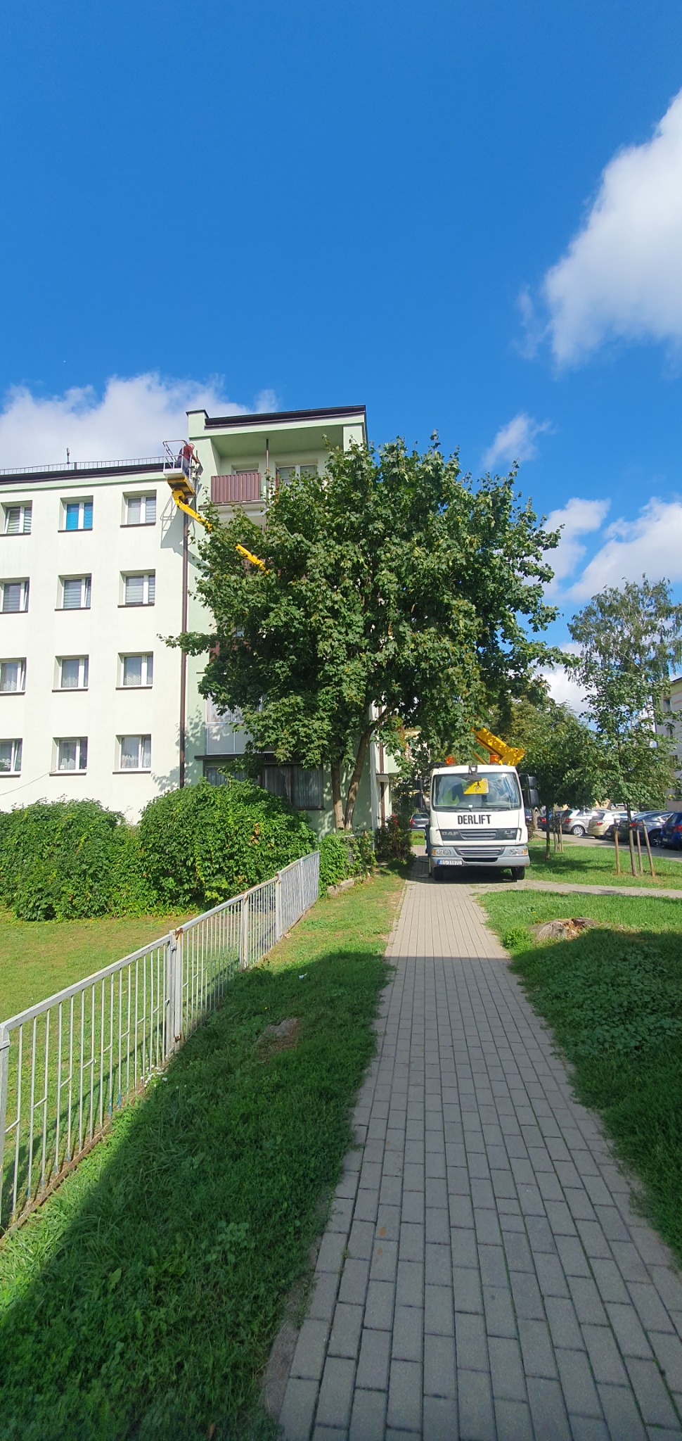 Biały budynek mieszkalny z żółtymi elementami podnośnika na tle błękitnego nieba z chmurami, widoczny z poziomu chodnika