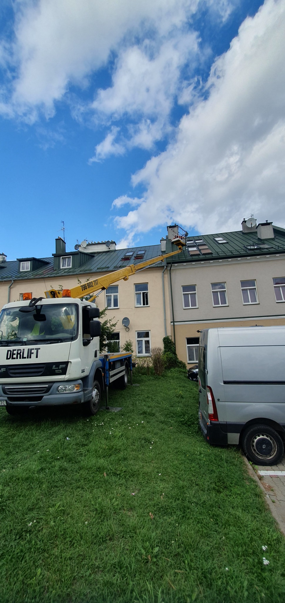 Żółty wysięgnik podnośnika firmy Derlift sięga dachu budynku z licznymi oknami dachowymi, na tle błękitnego nieba z obłokami.