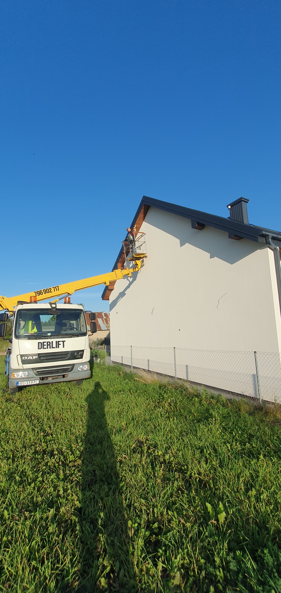 Pracownik na platformie żółtego podnośnika koszowego, wykonujący prace na wysokości przy ścianie domu jednorodzinnego z szarym dachem, widok z poziomu trawnika, cień osoby na pierwszym planie.
