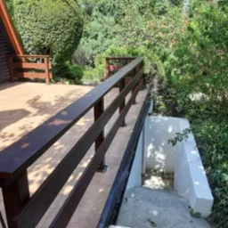 Zewnętrzny taras wyłożony płytkami z drewnianą balustradą, prowadzący do zejścia do piwnicy, otoczony zielenią i drzewami owocowymi.