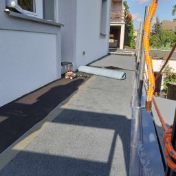 Piotr - Prace na dachu budynku: rozłożona papa bitumiczna, zwinięta rolka membrany dachowej i narzędzia dekarskie przy ścianie, widok z rusztowania.