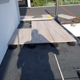 Piotr - Montaż tarasu na dachu: jasne płyty tarasowe ułożone na regulowanych wspornikach z tworzywa sztucznego na pokryciu bitumicznym, widoczny cień osoby i fragment miarki.