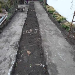 Piotr - Przygotowanie podłoża pod chodnik z wykopanymi rowami, widoczna taczka i osoba w tle, ogródek przed domem.