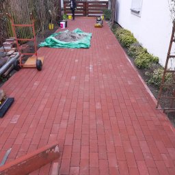 Piotr - Czerwona kostka brukowa ułożona na podwórku, widoczny wózek z narzędziami, ubijarka i osoba w oddali, ogród z elementami drewnianymi.