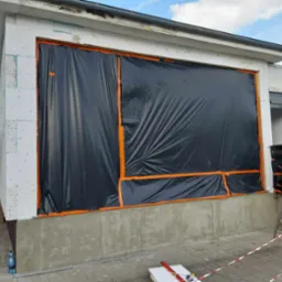 Budynek w trakcie prac: ocieplanie ścian zewnętrznych styropianem, zabezpieczone czarną folią z pomarańczową taśmą, widoczna drabina i butelka wody.